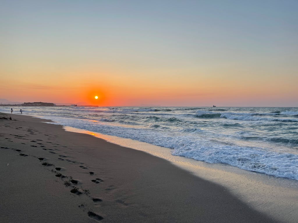 A tranquil beach sunset in Rethimno, Greece with footprints in the sand and gentle ocean waves.